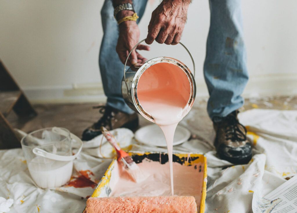 Person pouring light pink paint from a metal bucket into a yellow paint tray with a roller, preparing for interior wall painting in a home in Quebec or Montreal.
