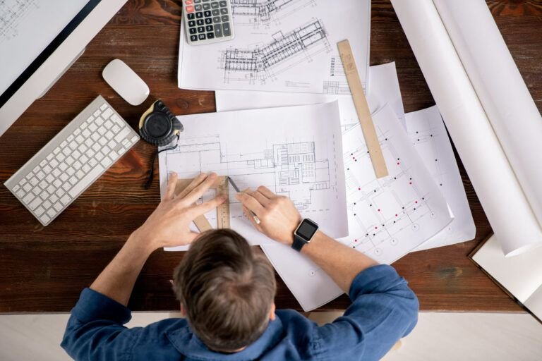 Architecture and engineering services Quebec showing architect or engineer working on building blueprints at desk with ruler calculator and computer in professional design workspace.