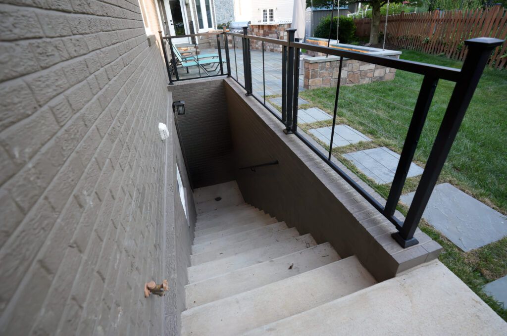 Basement Walkout Services in Montreal featuring an exterior concrete stairway leading to a lower-level entrance, with a safety railing, gray brick wall, and a terrace area with outdoor seating and a fire pit in the background.