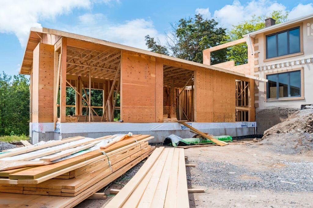 Home addition construction in Quebec showing a wood-frame extension built on a concrete foundation, with stacked lumber in front and weatherproof sheathing partially installed on the exterior wall.